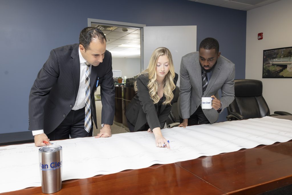 Three professionals in business attire lean over a large blueprint on a conference table, pointing and discussing.