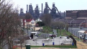 Skatepark with concrete bowls and skaters in the foreground, residential houses and a large industrial factory with smokestacks in the distance.
