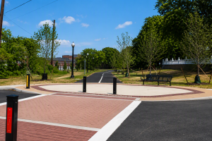 A circular brick plaza with a black bollard at the center, adjacent to a tree-lined path and a park bench under a bright blue sky.