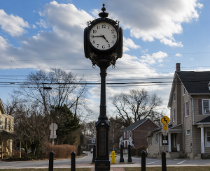 Ornate black clock tower in a small town square with leafless trees and residential houses in the background under a blue sky with clouds.