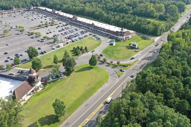 Aerial view of a large shopping plaza with pyramid-roof towers, long storefronts, and a expansive parking lot bordered by trees.