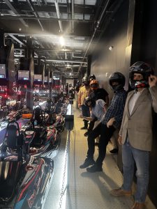 Indoor go-kart track pit row with multiple karts and drivers wearing helmets, some leaning on the railing along a metallic walkway.