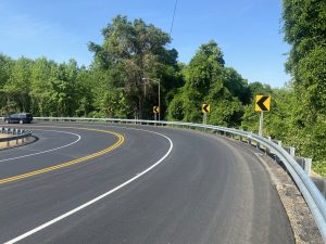 Curved highway with yellow chevron signs, guardrails, and dense green trees under a clear blue sky; a dark car on the left lane.