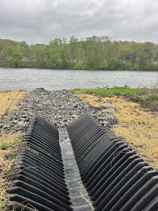 Black corrugated drainage culvert extending from a rocky path toward a lake with a tree-lined shore and cloudy sky in the background.