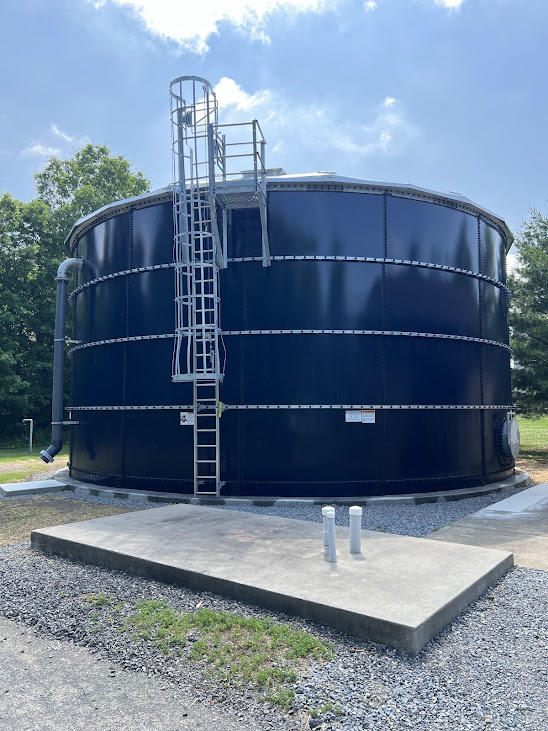 Large black cylindrical water storage tank with exterior metal ladder and piping, outdoors on a concrete pad under a blue sky.