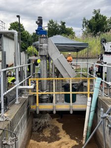 Vertical mixer on a platform over a brown wastewater basin at a treatment facility, with yellow railing and workers in the background.