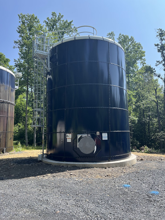 Large dark blue cylindrical storage tank with metal ladder and railing, outdoors among trees on a gravel site.