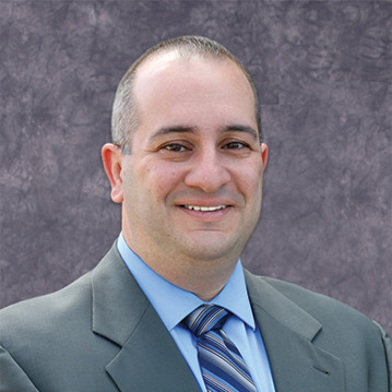 Portrait of a smiling man in a gray suit and blue striped tie against a textured gray backdrop, professional headshot.