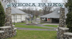 Stone entrance arch reads YEONA PARK with stone pillars; beyond are paved paths, a circular fountain with a clock, and park buildings.