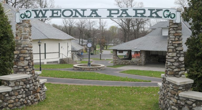 Stone entrance arch reads YEONA PARK with stone pillars; beyond are paved paths, a circular fountain with a clock, and park buildings.