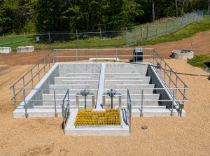 Rectangular concrete water-treatment basin with dividing walls and surrounding metal railings, a bright yellow grate in the foreground.