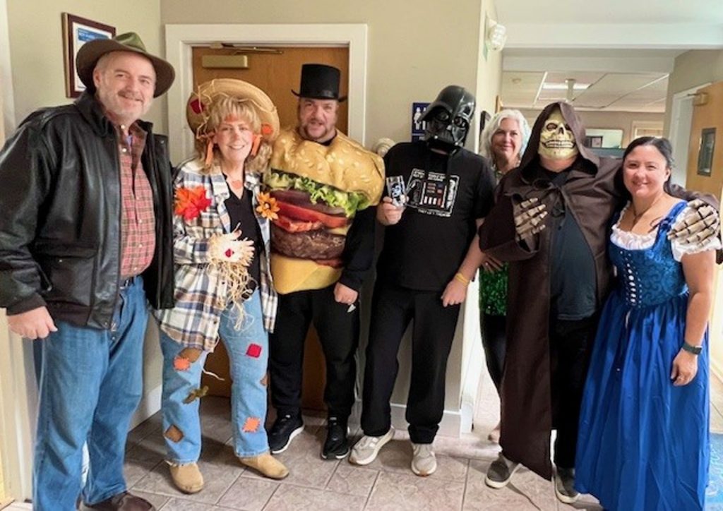 Group of people in costumes posing together in a hallway, including a giant hamburger, Darth Vader, a skeleton, and a blue-dirndl dress wearer.