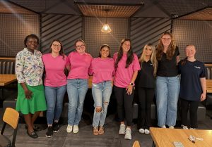 Eight women standing together in a cafe, smiling at the camera; some wearing pink shirts and jeans, others in black tops, floral blouse or navy shirt in a casual group photo.