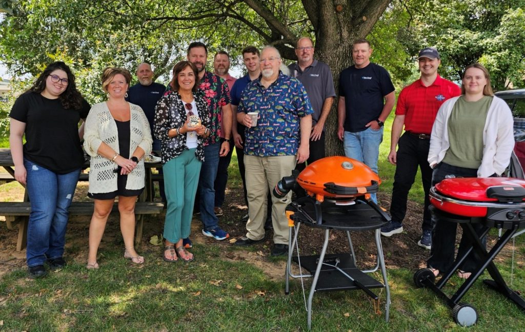 Group of adults posing for a photo at an outdoor company barbecue under a tree, with two portable grills in the foreground.