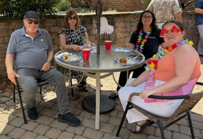 Four adults seated around a metal patio table outdoors, with plates and red cups, wearing leis and casual clothes.