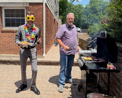 Two men barbecuing outdoors; one wears sunflower sunglasses and a lei, holding a platter, while the other stands by a charcoal grill with food trays near a brick building.