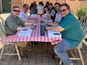 Group of people seated around a long outdoor table with a red-and-white checkered tablecloth, smiling and chatting on a sunny day in a brick courtyard.