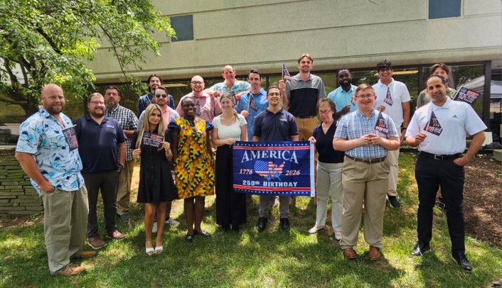 Group of diverse adults posing outdoors, holding a banner celebrating America’s 250th birthday (1776–2026).