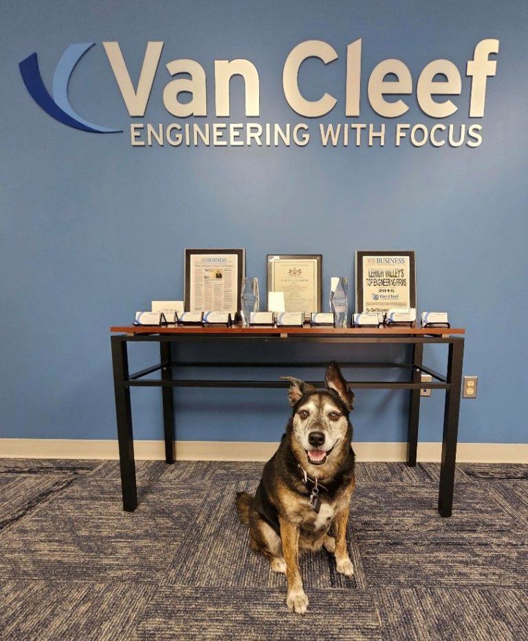 Dog sitting in front of a reception table with awards and the Van Cleef Engineering sign on a blue wall.