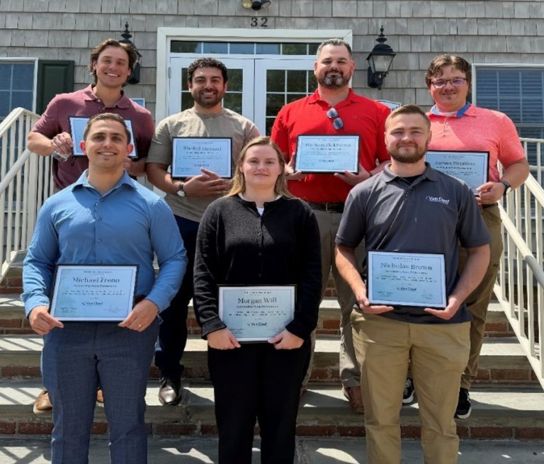 Group of eight adults on steps, each holding a certificate or plaque for an achievement outside a gray building with the number 32 above the door.