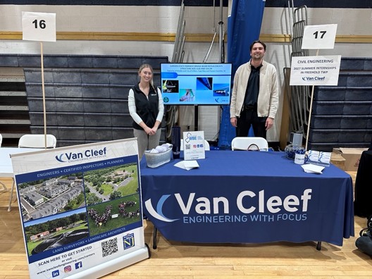 Engineering booth for Van Cleef with two presenters behind a blue tablecloth-covered table in a gymnasium display area, signs 16 and 17 visible.