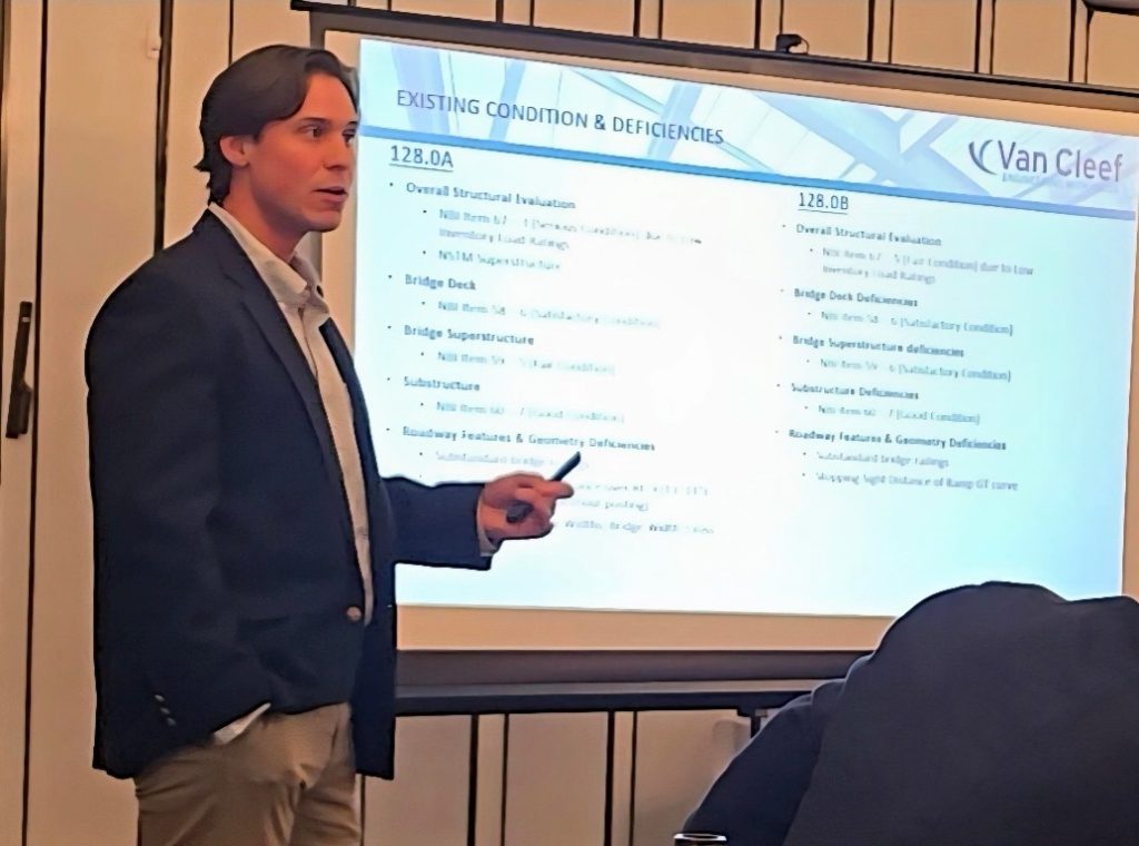 Business presenter in a blazer points to a projected slide titled 'Existing Conditions & Deficiencies' during a meeting.