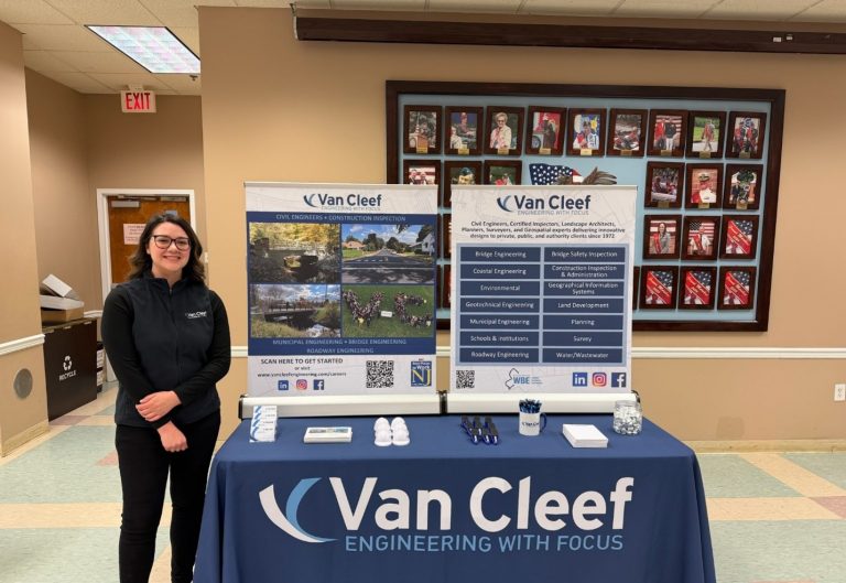 Woman in a dark vest stands beside a Van Cleef Engineering booth with two vertical banners and a staffed table in a hallway.