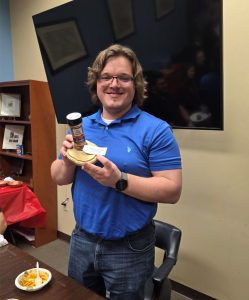Man in a blue polo shirt holding a wooden award base with a spice grinder and a note, smiling at the camera.