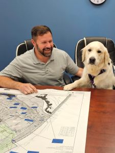 Man and golden retriever seated at a conference table with a large map spread out, both looking pleased in a planning setting.