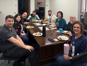 Nine adults sit around a long conference table eating pizza and wings in a casual office setting, with drinks and a pink tumbler visible.