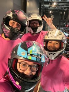 Four people wearing pink shirts and full-face motorcycle helmets take a group selfie, smiling at the camera indoors.