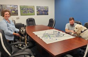 Woman in a denim shirt sits at a conference table with a small black-and-tan dog; a man with a light-colored retriever sits on the other side as a large map lies on the table.
