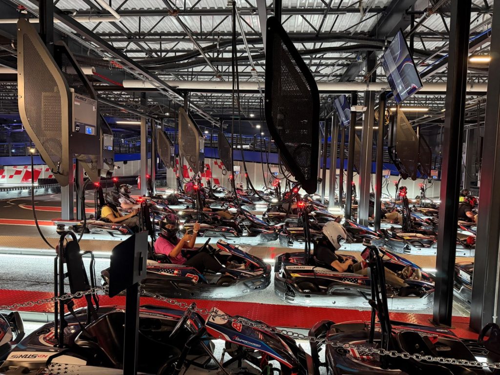 Indoor go-kart arena with multiple drivers seated in their karts at the starting grid, wearing helmets and ready to race.