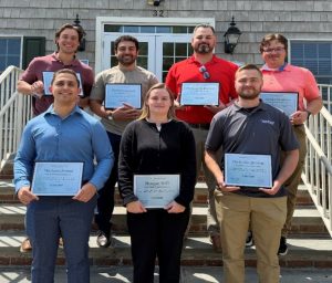 Eight adults pose on outdoor steps, each holding a certificate or plaque, facing the camera.