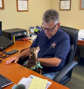 Man in a navy polo shirt sits at a cluttered office desk, a small brown dog in a teal harness looks up at him affectionately from his lap.