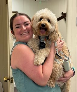 Woman in a teal sleeveless top hugs a fluffy beige dog with a harness, both smiling at the camera indoors.