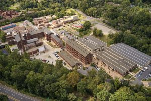Aerial view of a brick factory campus with connected buildings, a tall smokestack, loading areas, and surrounding trees.