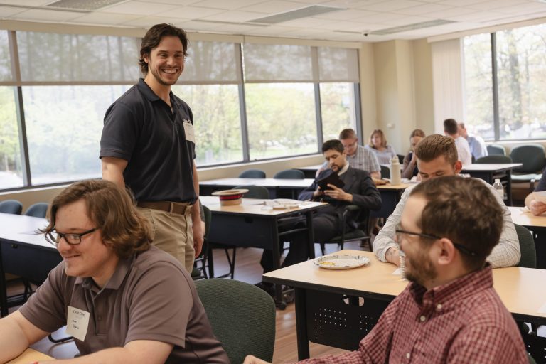 Smiling man in a dark polo shirt stands at the front of a classroom while others sit at tables with plates and cups.