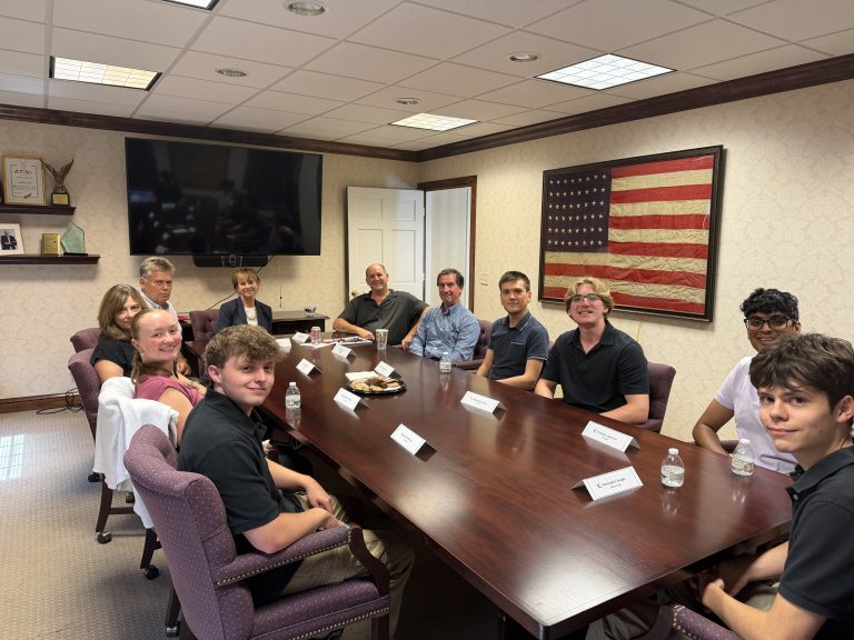 Group of people seated around a large conference table in a meeting room, with an American flag on the wall and name cards, snacks, and water bottles on the table.