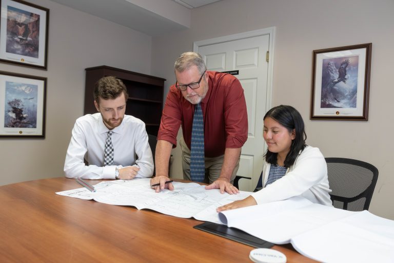 Three colleagues review architectural plans on a conference table, one man in a red shirt points to the blueprint.