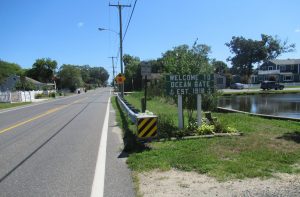 'Welcome to Ocean Gate' sign by a canal with houses along a sunny street; est. 1918 is on the sign text included in the image