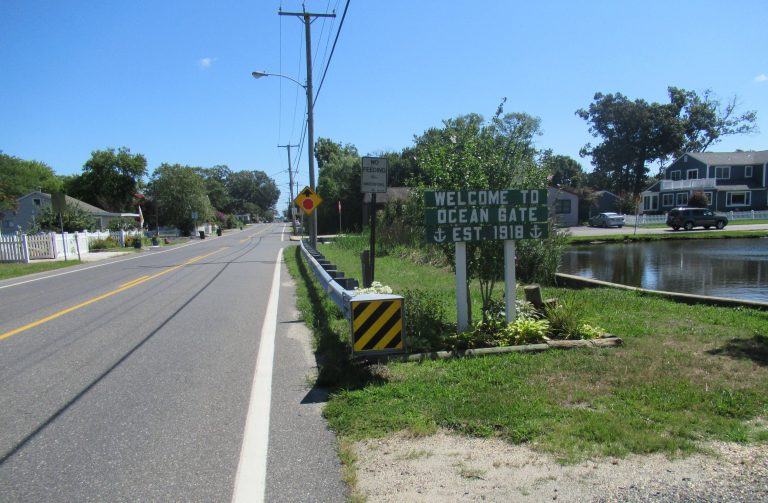 'Welcome to Ocean Gate' sign by a canal with houses along a sunny street; est. 1918 is on the sign text included in the image
