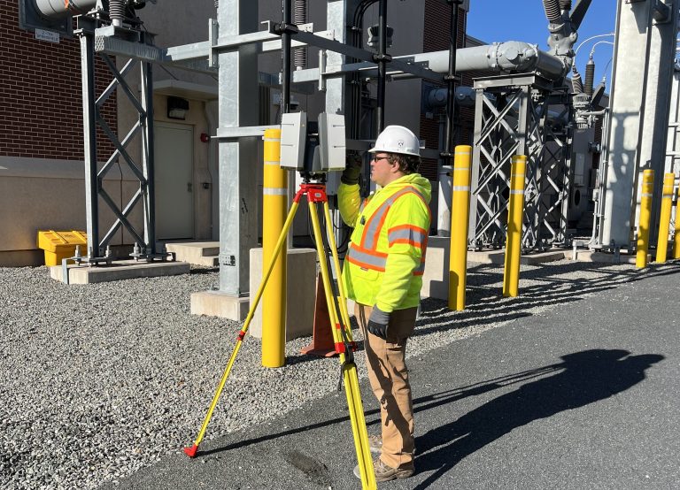 Utility worker in a neon safety jacket and hard hat using a tripod surveying instrument beside industrial equipment.